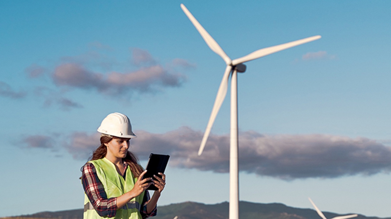 Una mujer con chaleco de seguridad amarillo y casco blanco utiliza una tablet para controlar un dron delante de un aerogenerador.