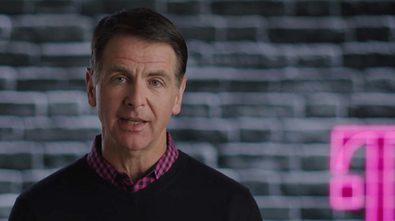 Hombre con camisa a cuadros magenta y negra y suéter negro delante de una pared de ladrillos gris