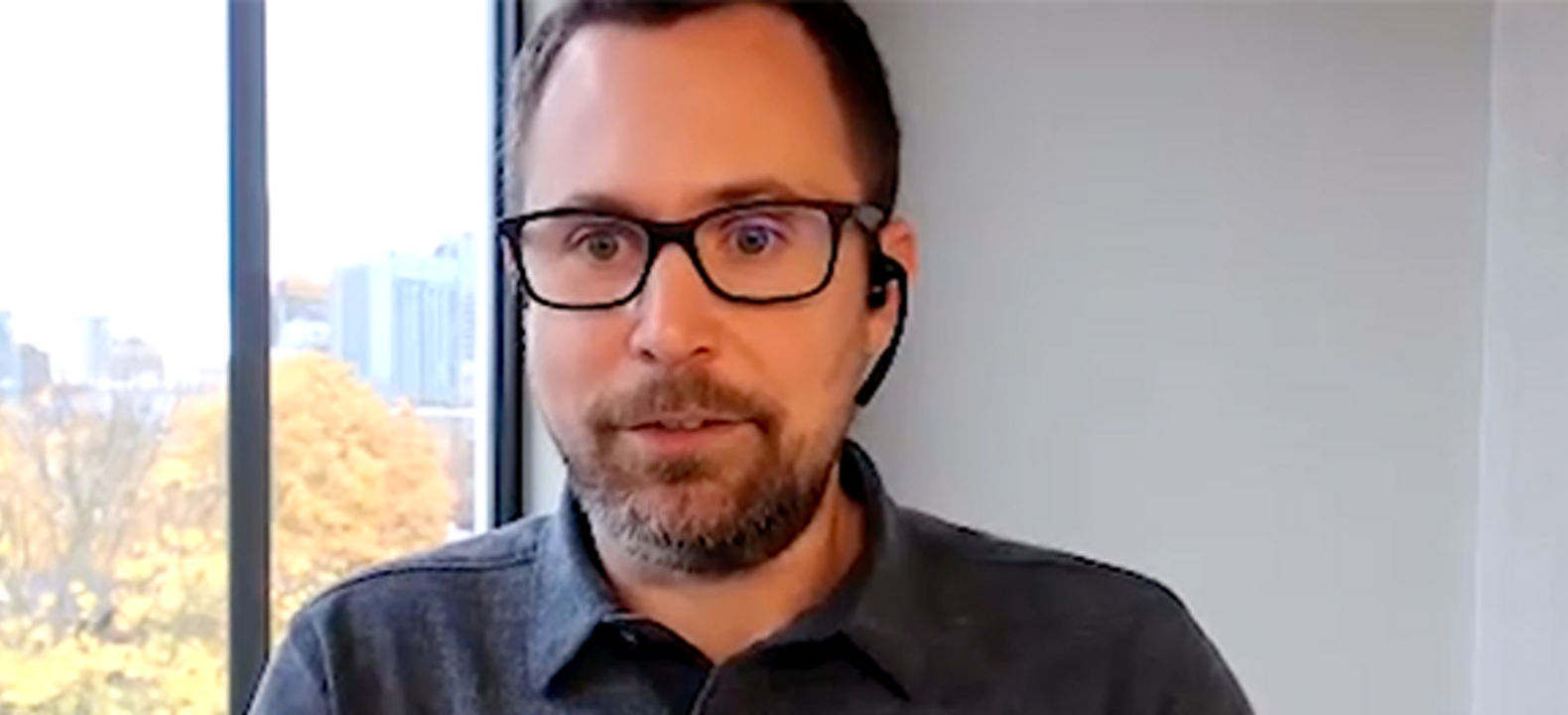 Primer plano de un hombre con barba, bigotes, gafas y una camisa negra con botones, con una ventana al costado