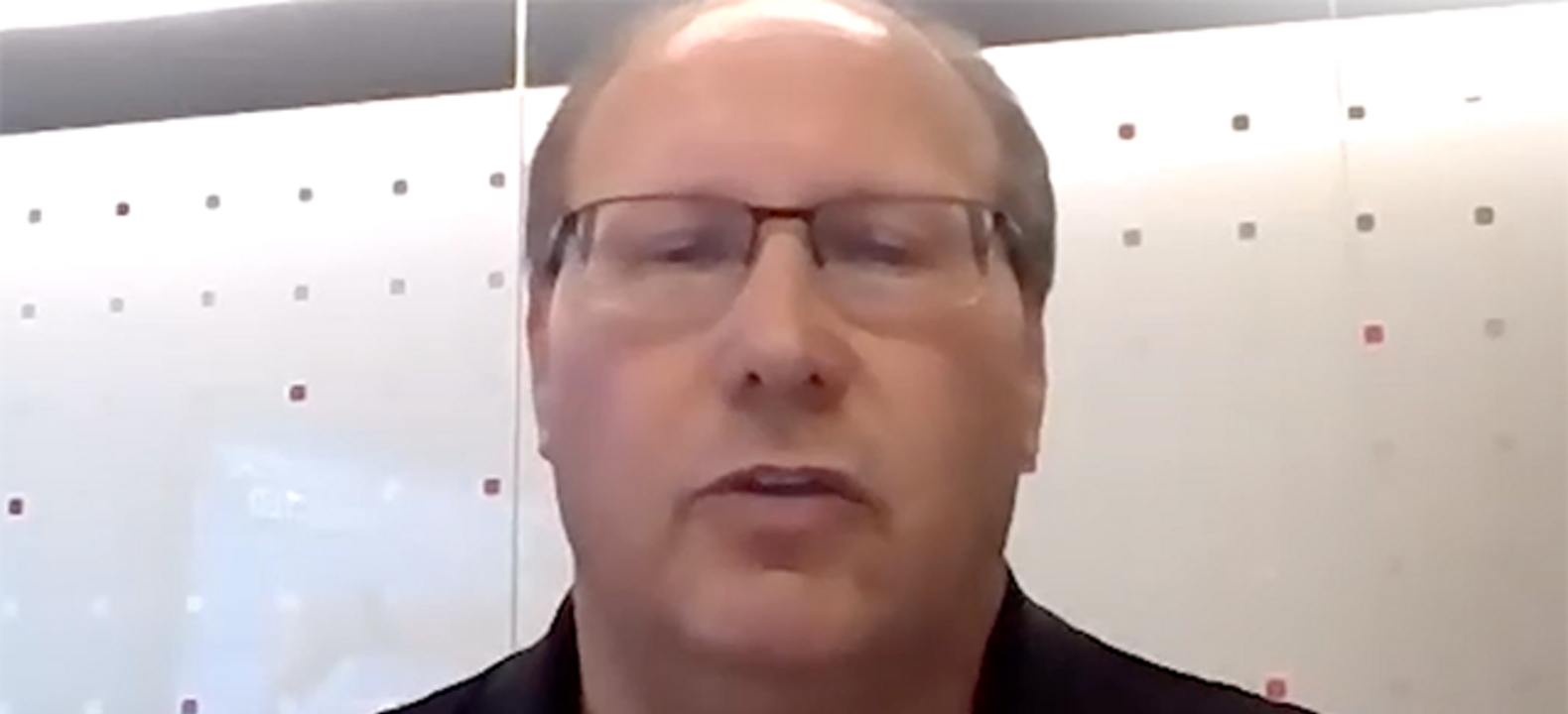 Primer plano de un hombre calvo con gafas y una camisa oscura sentado delante de un fondo blanco