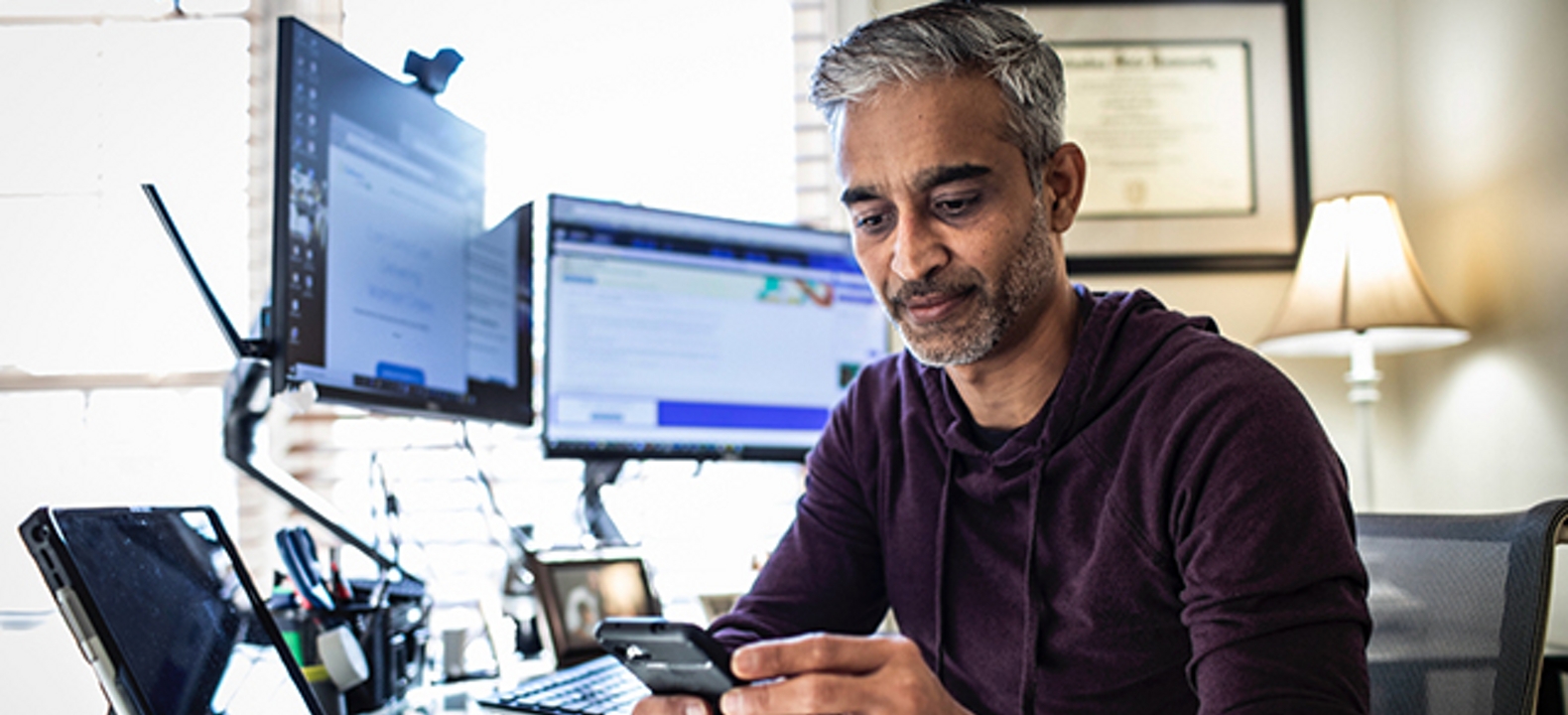 Hombre sentado en una oficina en el hogar mirando un celular junto a múltiples pantallas de computadora.