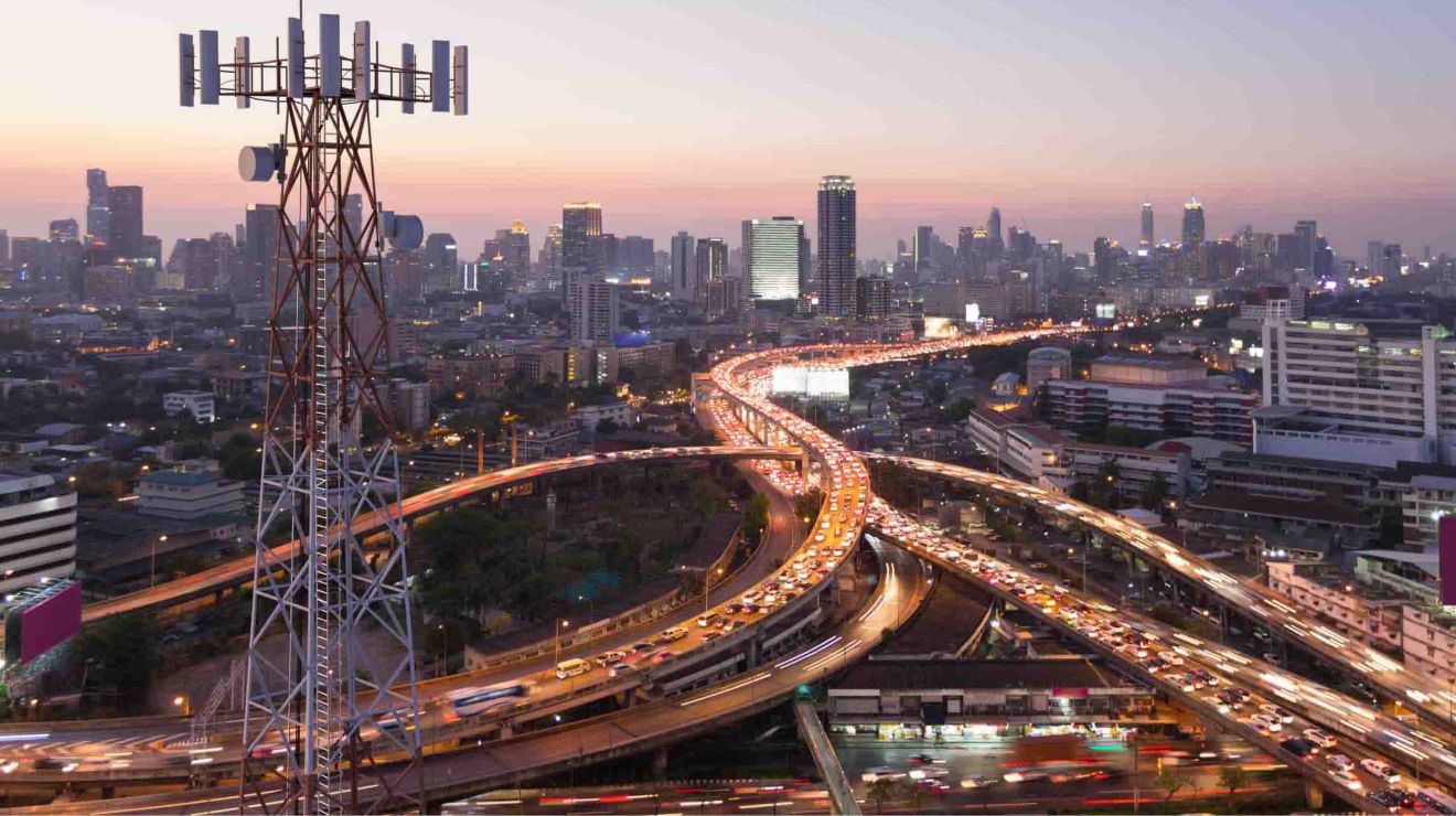 Vista aérea de autopistas muy transitadas con una torre de telefonía móvil en primer plano y una ciudad a lo lejos.