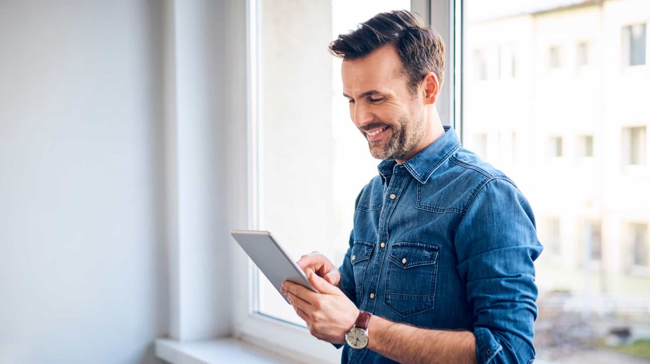 Profesional de los negocios sonriente, reclinado contra la ventana de una oficina, escribe en una tablet conectada.