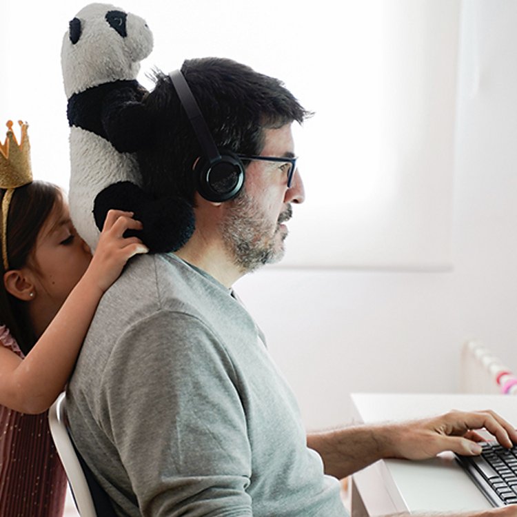 Hombre sentado ante una computadora con los audífonos puestos mientras una niña sostiene un oso panda sobre su cuello.