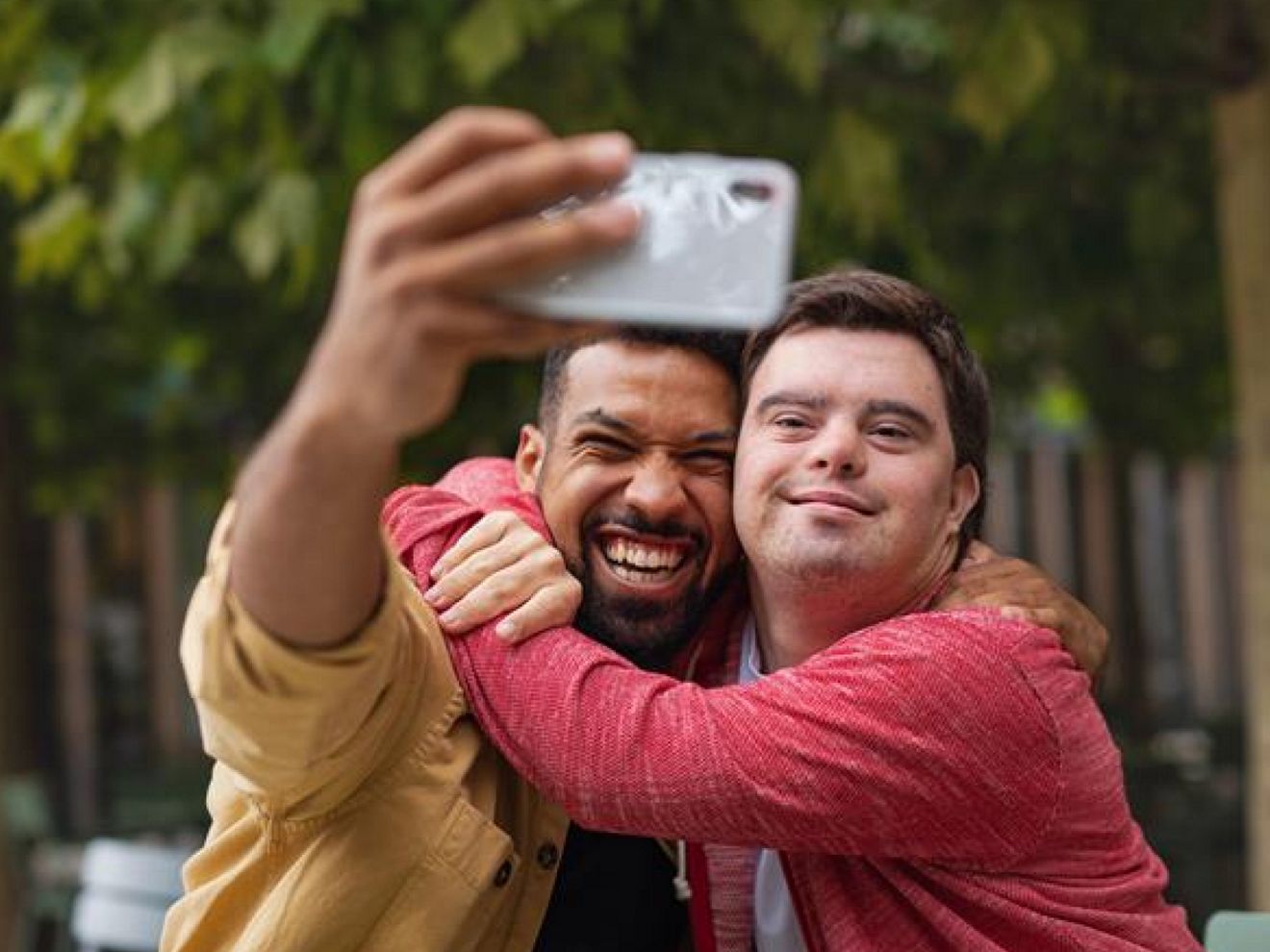 Dos amigos ríen mientras se toman un selfie juntos en un jardín iluminado por el sol.