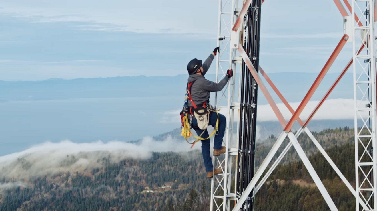 Un trabajador de torre con equipo de seguridad sube a una torre celular en una zona rural.