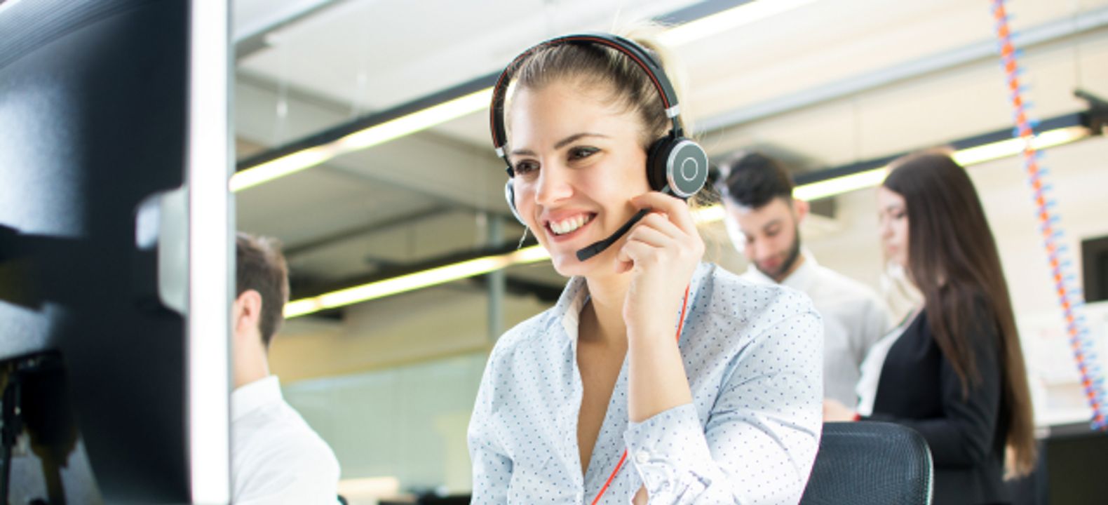 Una profesional de ventas sonriendo durante una llamada en la oficina; tres colegas están parados en el fondo.
