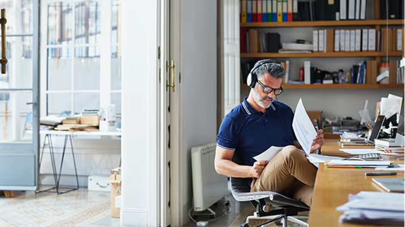 Hombre trabajando en casa con audífonos mientras mira papeles en su regazo.