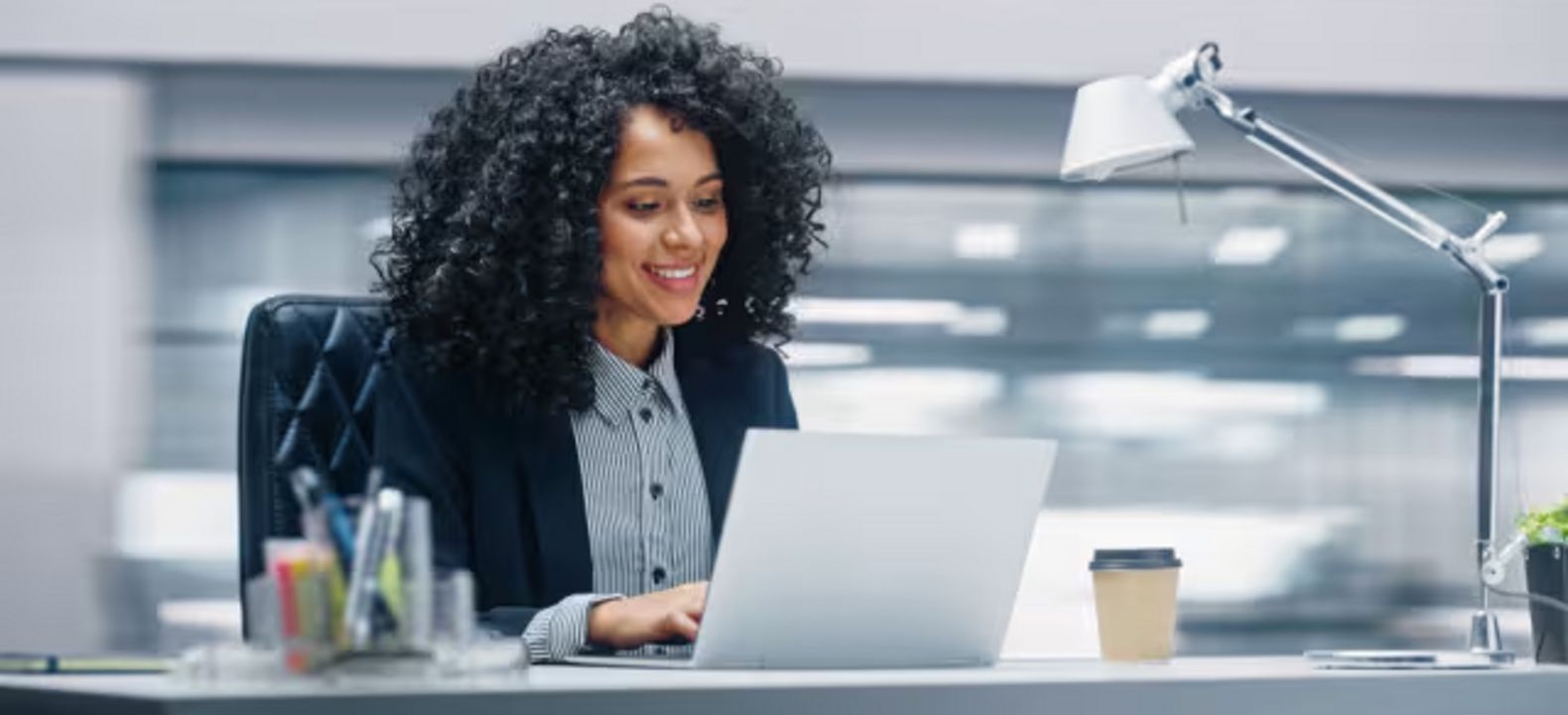 Mujer de negocios sonríe sentada a su escritorio mientras lee en su laptop.