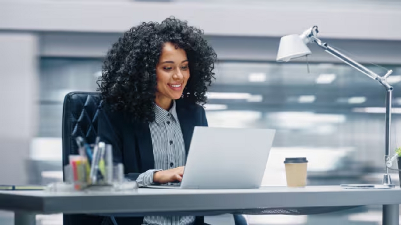 Mujer de negocios sonríe sentada a su escritorio mientras lee en su laptop.