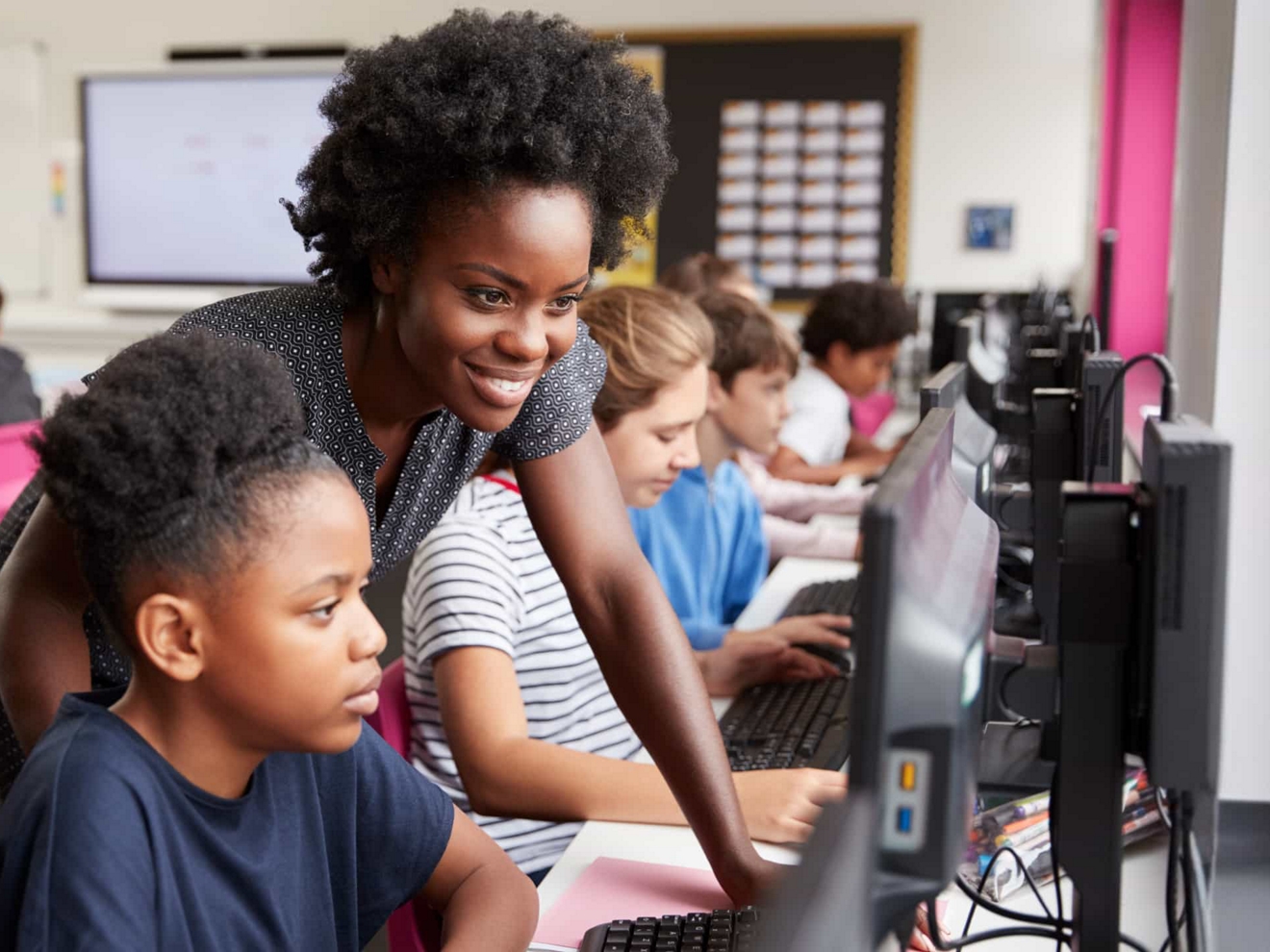 Profesora y una alumna mirando una computadora en un aula llena de gente.