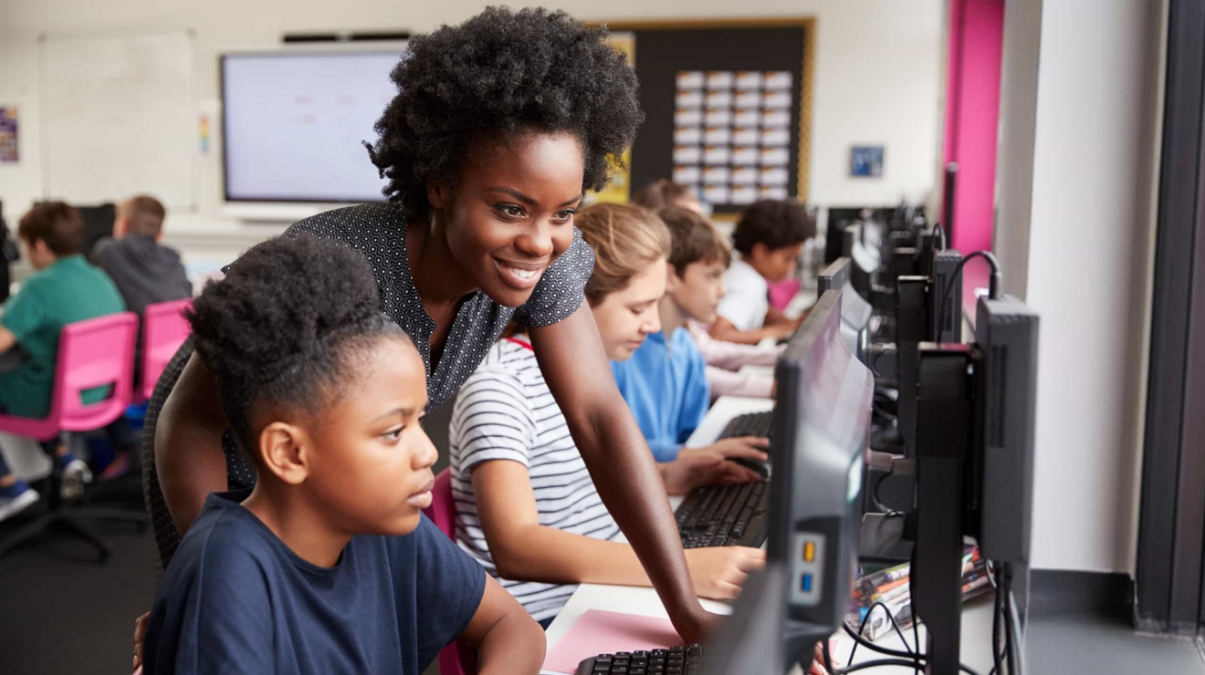 Estudiantes trabajan de forma independiente en computadoras de escritorio mientras un profesor revisa su trabajo.