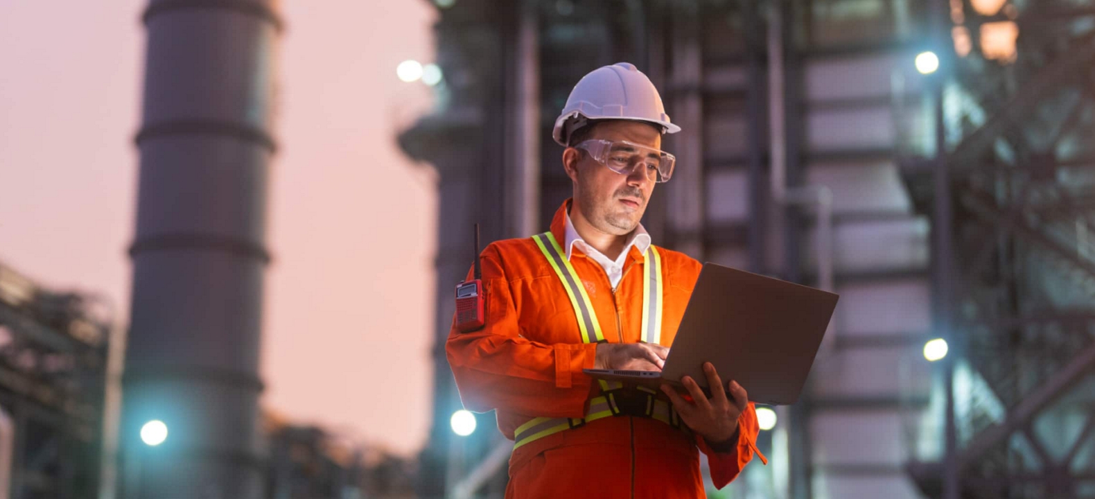 Un ingeniero en una refinería de petróleo estudia su tablet mientras está parado en el exterior al anochecer.