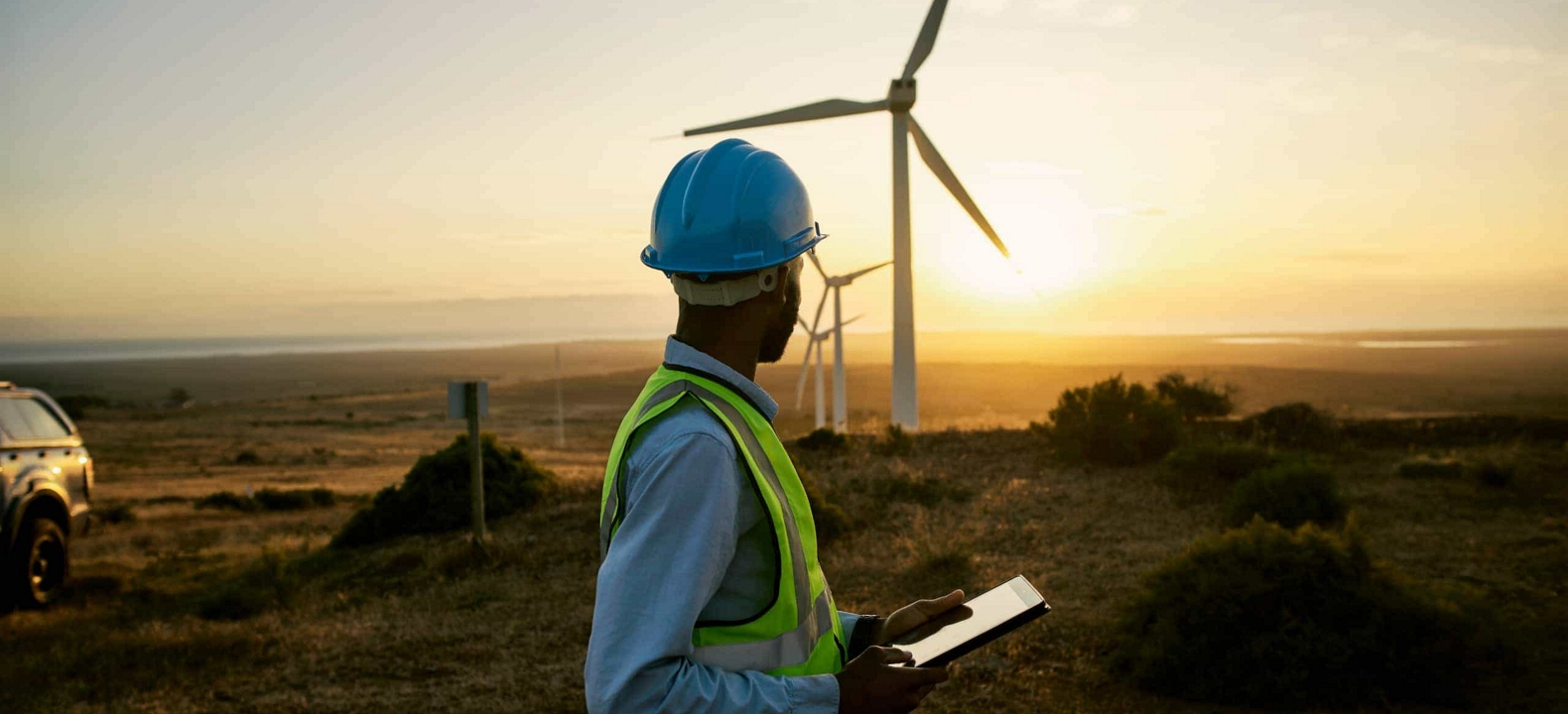 Un ingeniero mira hacia arriba sosteniendo su tablet para observar una fila de turbinas eólicas con la puesta de sol.