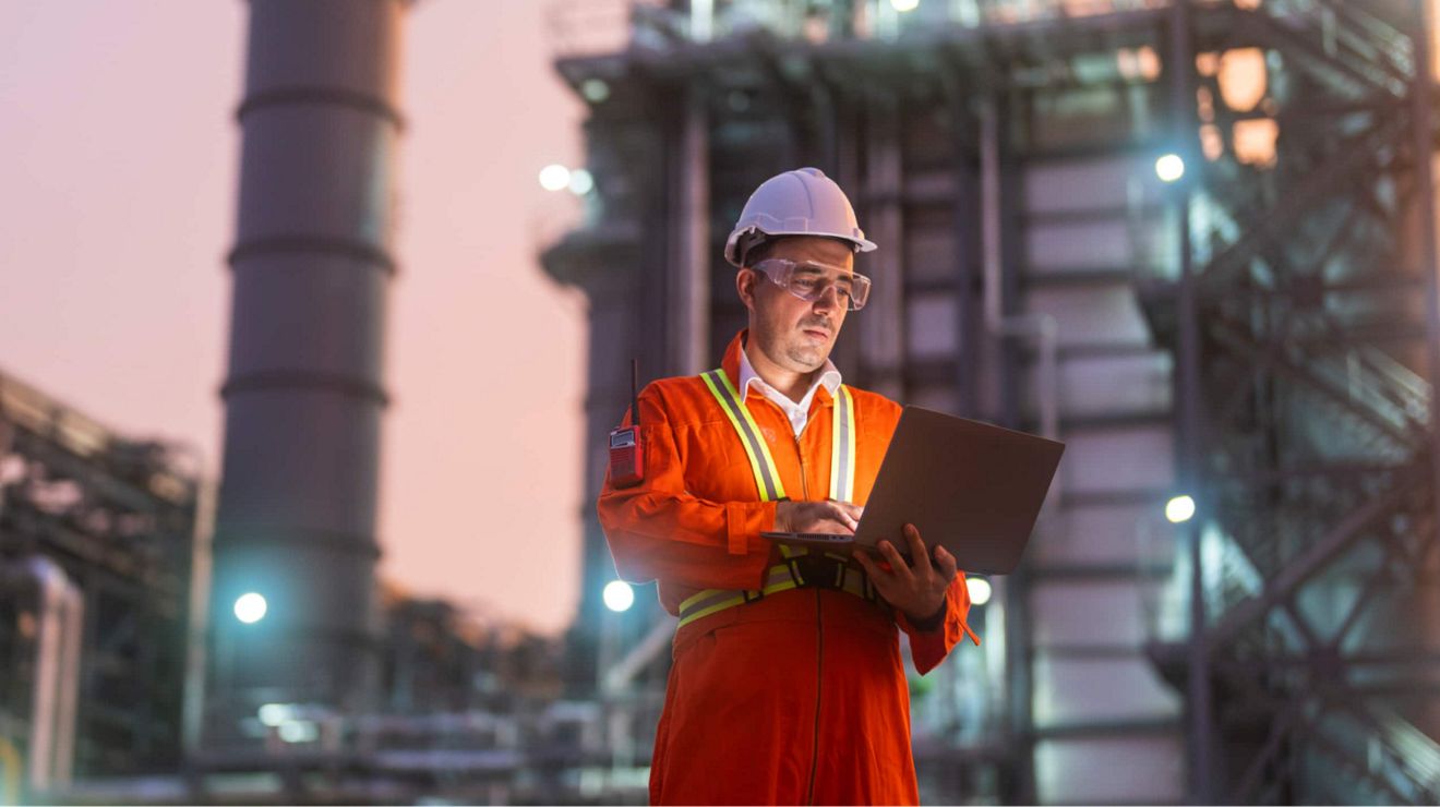 Un ingeniero en una refinería de petróleo estudia su tablet mientras está parado en el exterior al anochecer.