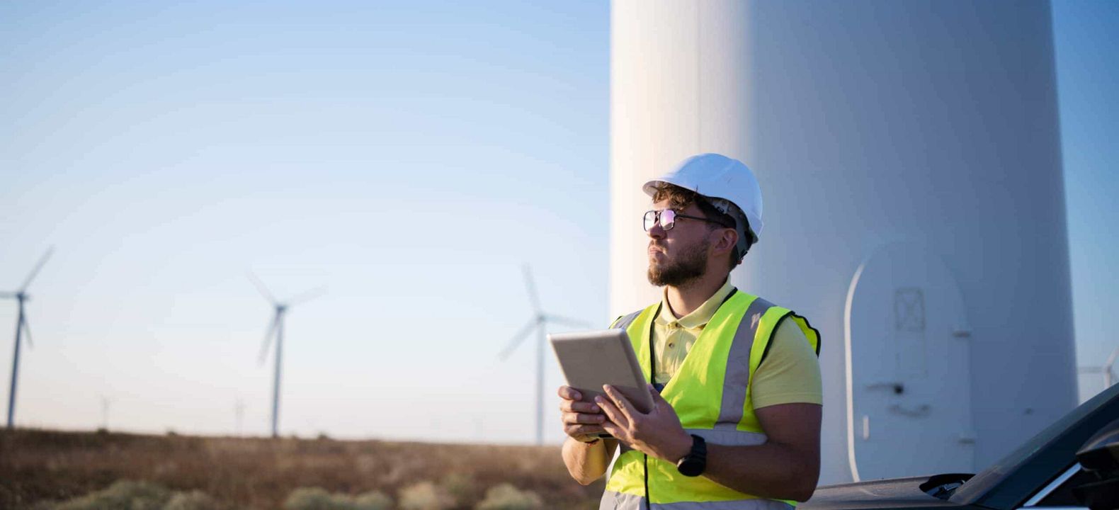 Un trabajador de servicios públicos en una granja eólica observa el horizonte con su dispositivo con conexión 5G en la mano.