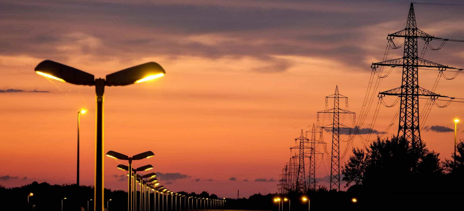 Una fila de torres eléctricas de alto voltaje junto a una fila de luces de la calle encendidas al anochecer.