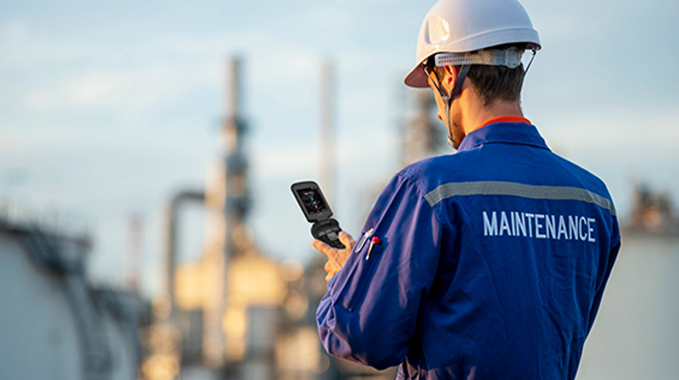 Un trabajador de mantenimiento revisa su teléfono móvil, con una refinería de petróleo a lo lejos.