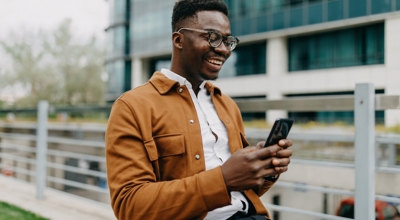 Un profesional de servicios financieros sonriente utiliza su smartphone mientras camina por un campus empresarial.