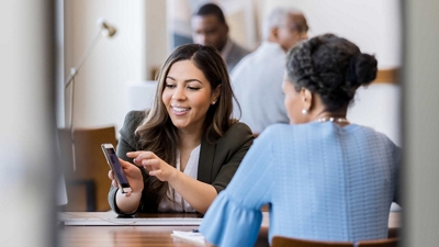 Una profesional de las finanzas muestra la pantalla de su dispositivo móvil a una colega al otro lado de la mesa.