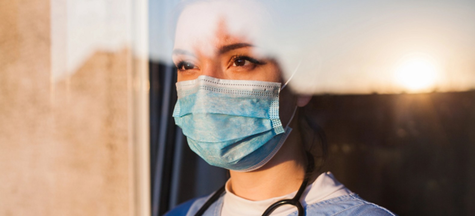 Una profesional médica con mascarilla mira a lo lejos a través de una ventana.