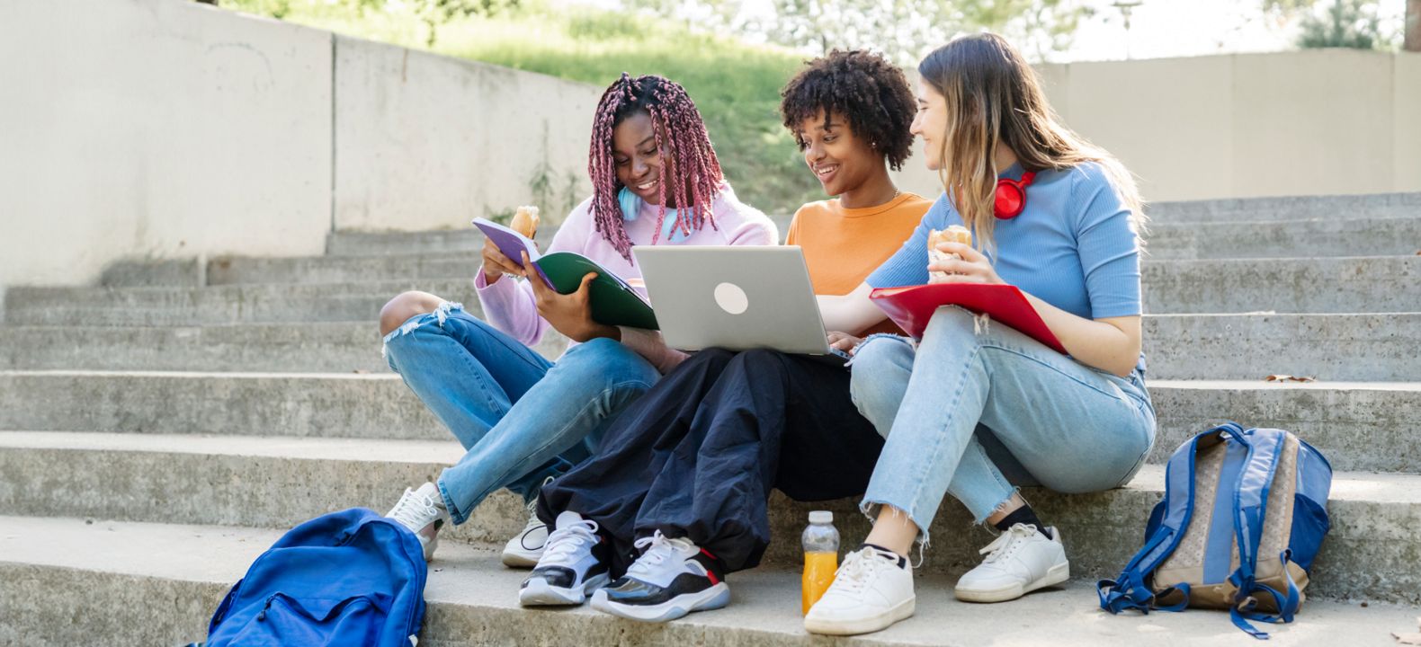 Tres estudiantes están sentados juntos en los amplios escalones del campus bajo el sol y comparten una laptop conectada.