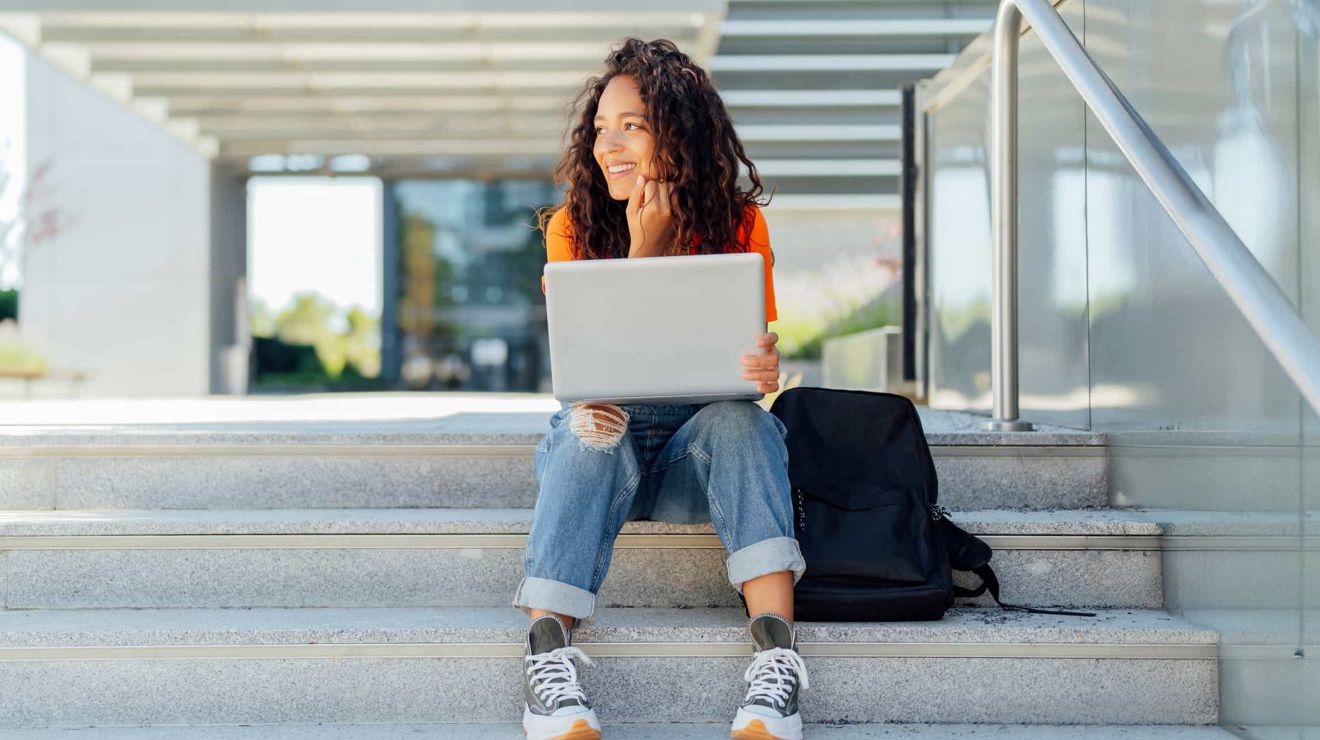 Un estudiante universitario feliz sentado frente a un edificio del campus con una laptop abierta sobre las rodillas.