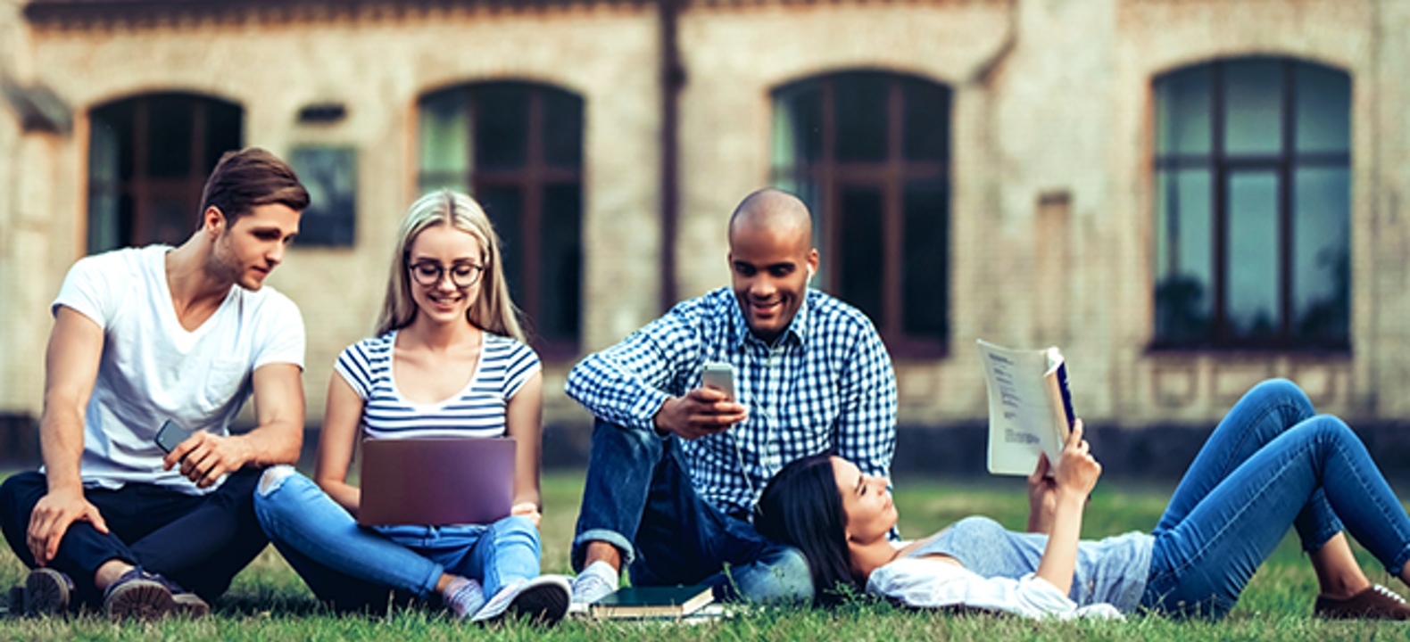 4 estudiantes en el césped del campus mirando sus dispositivos y apuntes
