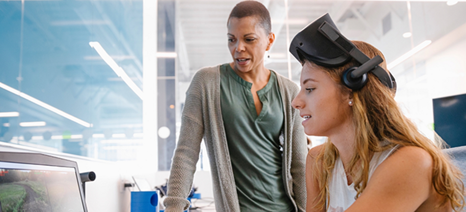Una estudiante trabaja frente a un monitor con unas gafas de RV sobre la cabeza mientras una profesora observa.