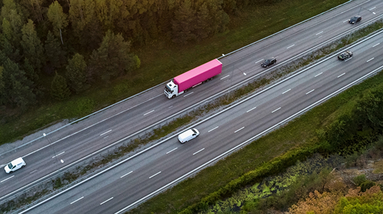 Vista aérea de una carretera con una camioneta blanca y un camión grande en color magenta y blanco