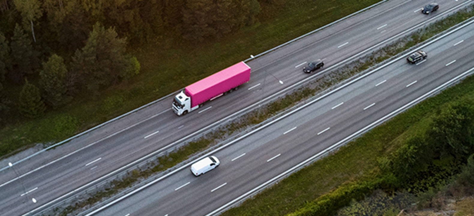 Vista aérea de una carretera con una camioneta blanca y un camión grande en color magenta y blanco