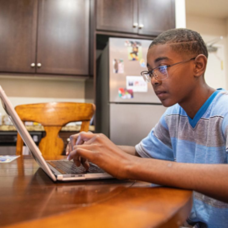 Niño con gafas y camiseta azul y gris sentado en la cocina trabajando con una laptop.
