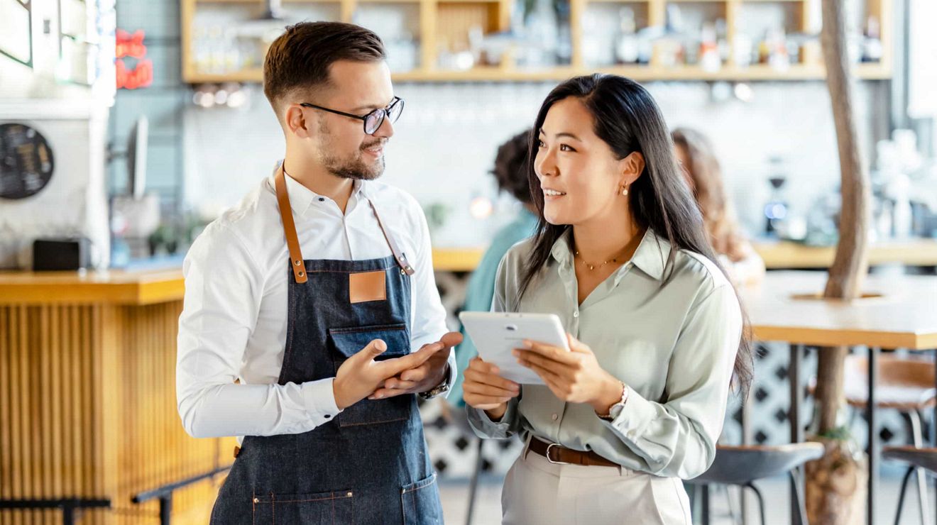 Propietario de pequeños negocios y un cliente revisan información en una tablet dentro de una cafetería.