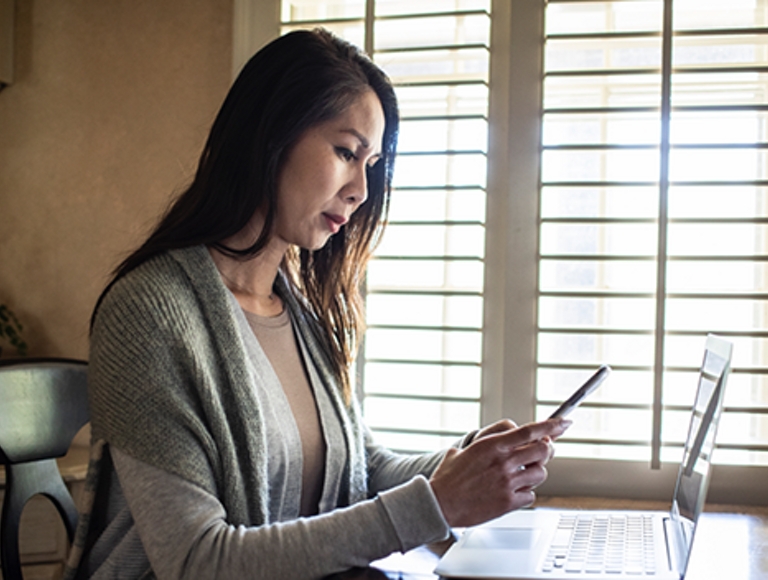 Mujer sentada a la mesa en su casa, cerca de una ventana, con una laptop delante de ella mientras mira su smartphone
