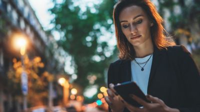 Una profesional concentrada revisa su smartphone mientras camina por la ciudad al atardecer.