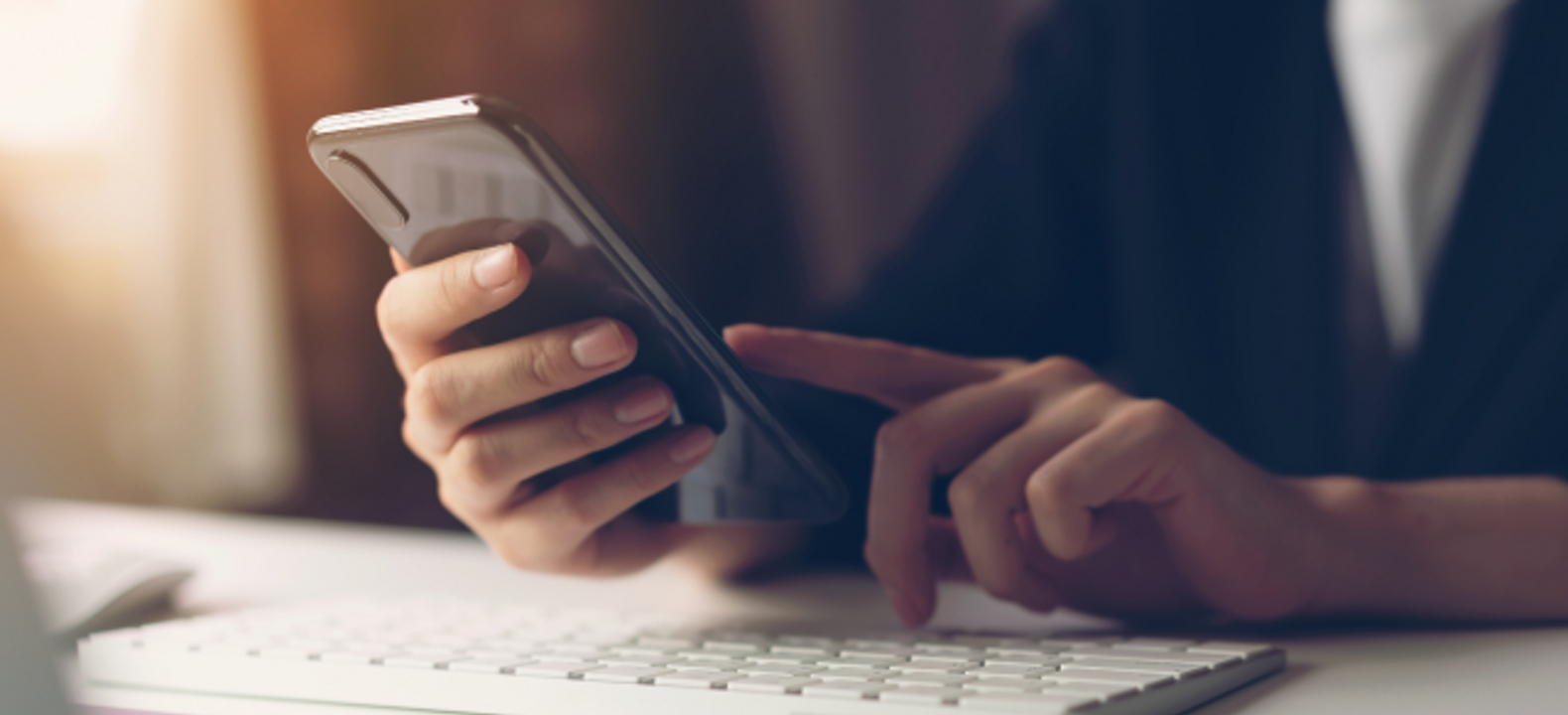 Primer plano de dos manos usando un smartphone sobre el teclado de una computadora.