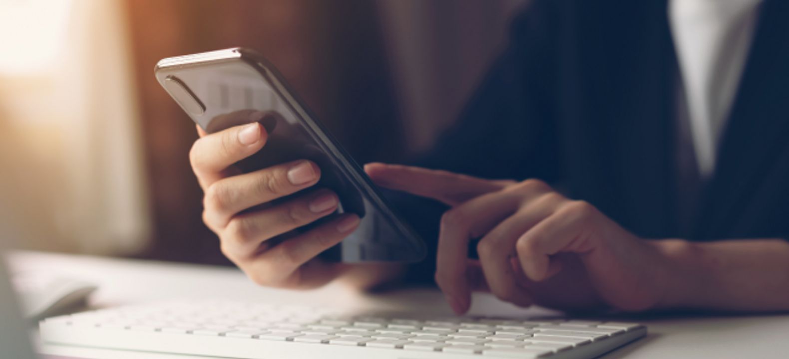 Primer plano de dos manos usando un smartphone sobre el teclado de una computadora.