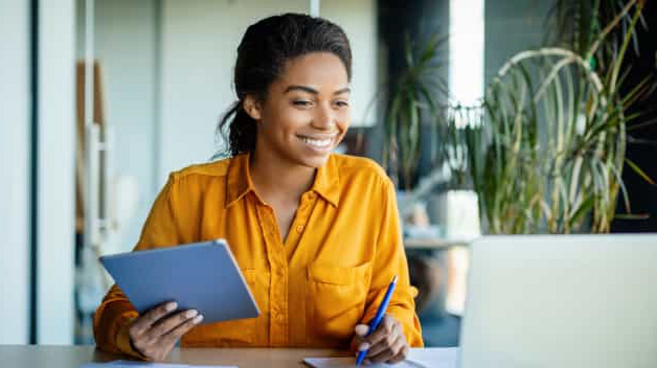 Una profesional sonríe mientras sostiene una tablet y toma notas durante una videollamada en su laptop.
