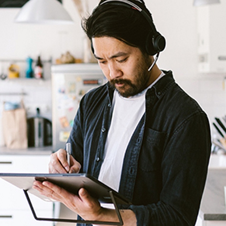 Un joven con audífonos parado en la cocina mientras escribe en su tablet.