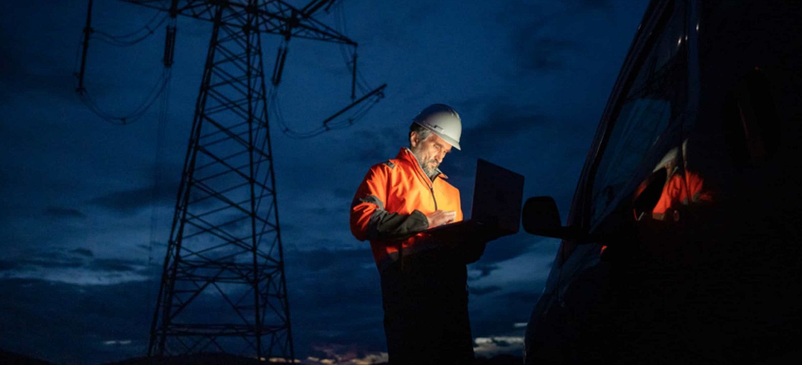 Al atardecer, un técnico se encuentra fuera de su vehículo y trabaja con una laptop bajo una torre de alta tensión.