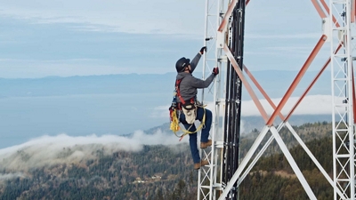 A tower worker wearing safety gear climbs a cell tower in a rural location