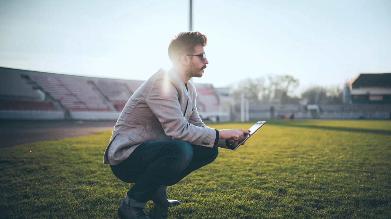 Un entrenador de equipo está agachado en el campo de juego, escribiendo notas en su tablet.