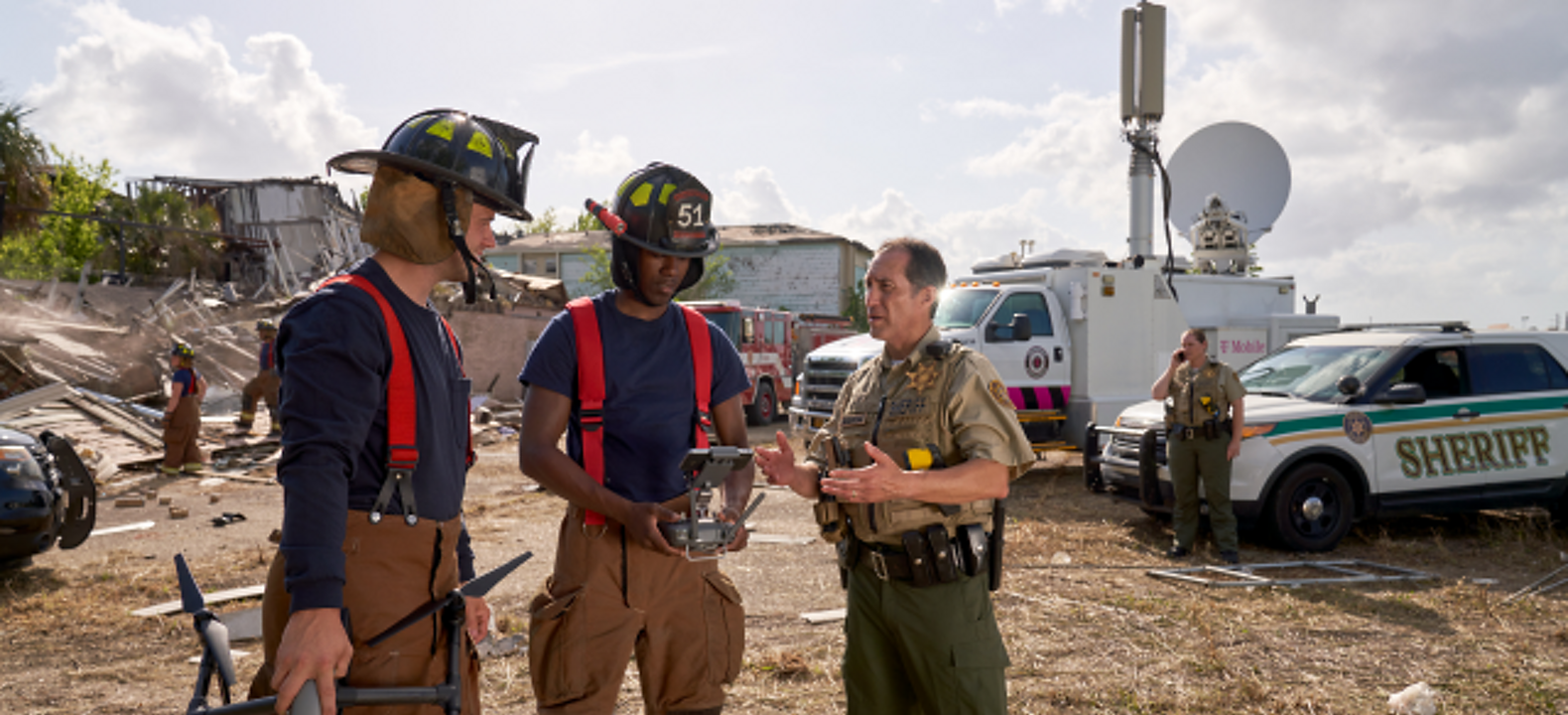 Dos bomberos sostienen un dron y su control mientras hablan con un alguacil en un sitio de respuesta a una emergencia.