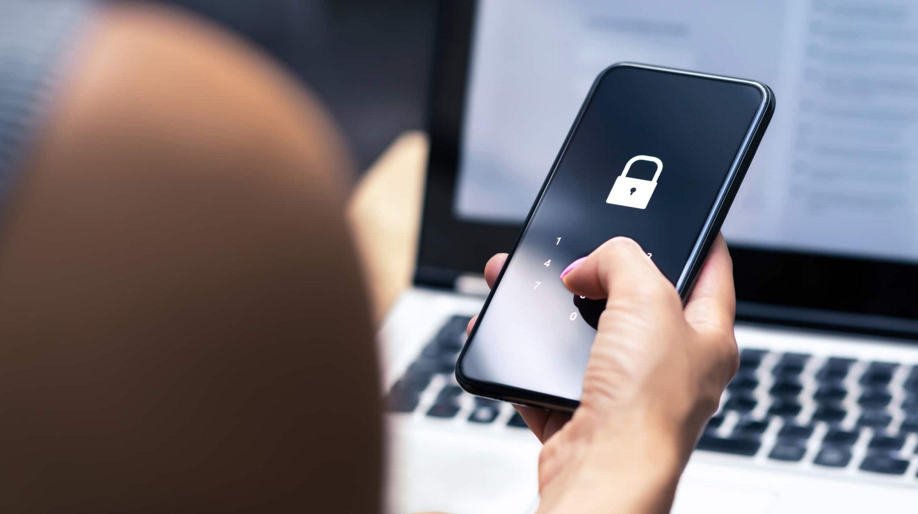 Vista por encima del hombro de la pantalla de bloqueo de un smartphone y una laptop al fondo.
