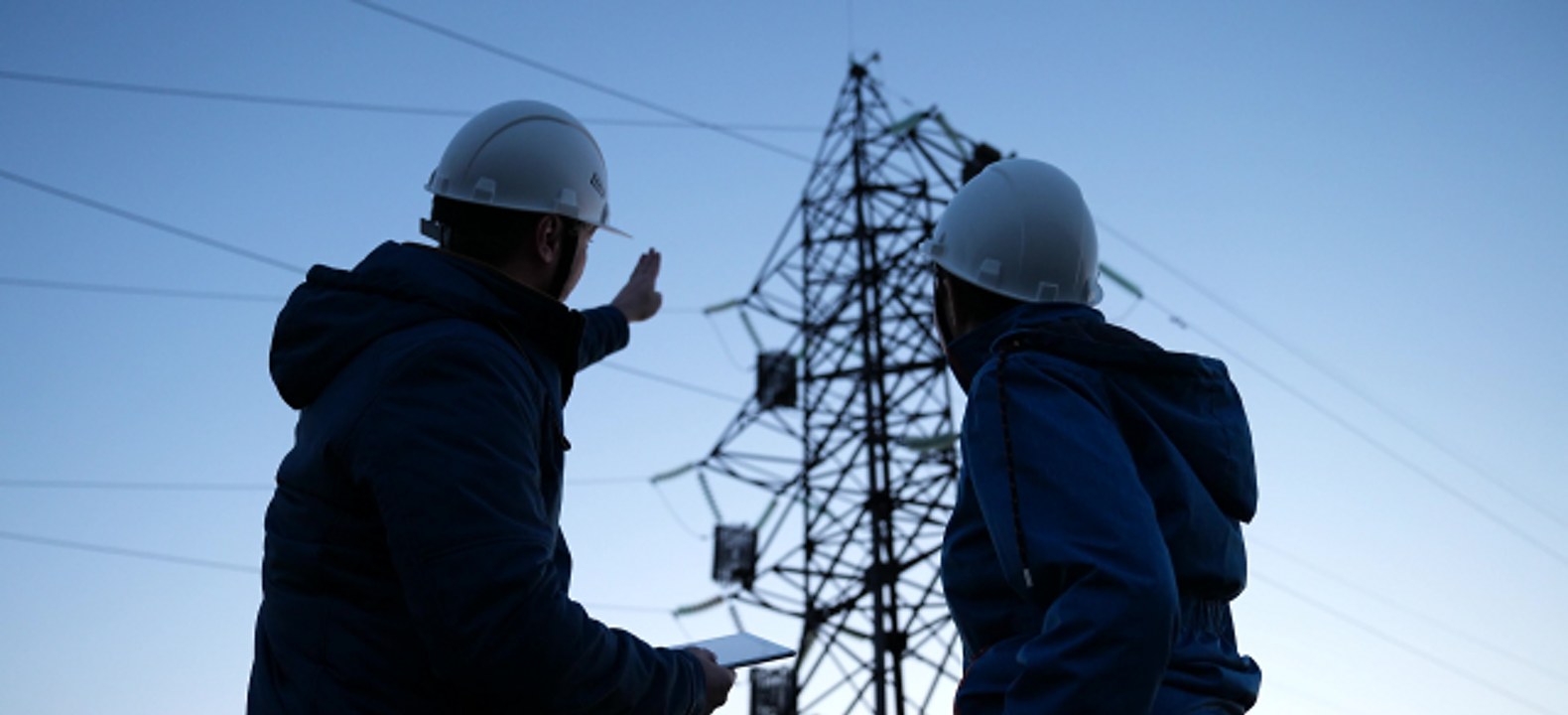 Dos técnicos del sector energético utilizando una tablet mientras evalúan una torre de tendido eléctrico.