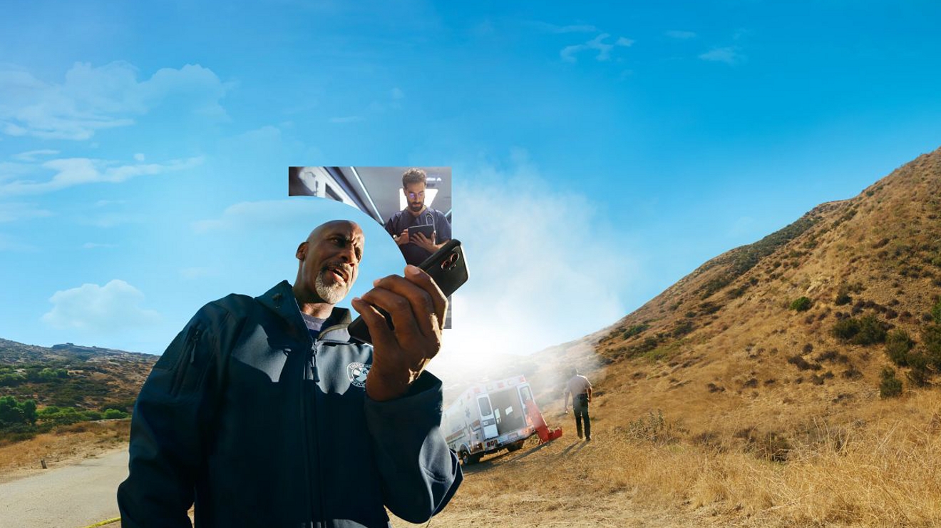 Un socorrista sostiene un teléfono cerca de una ambulancia al aire libre, bajo un cielo azul claro.