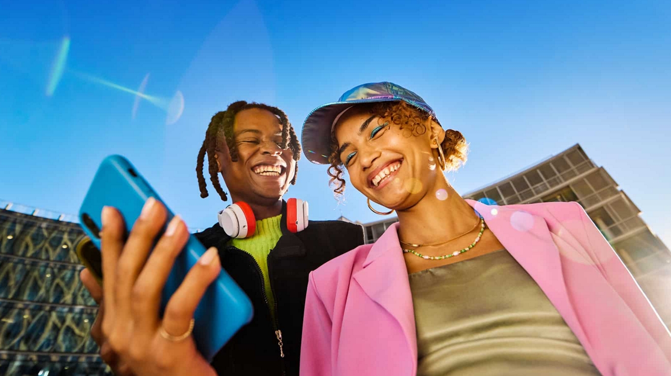 Pareja riendo mientras mira un dispositivo, con edificio y cielo azul en el fondo.