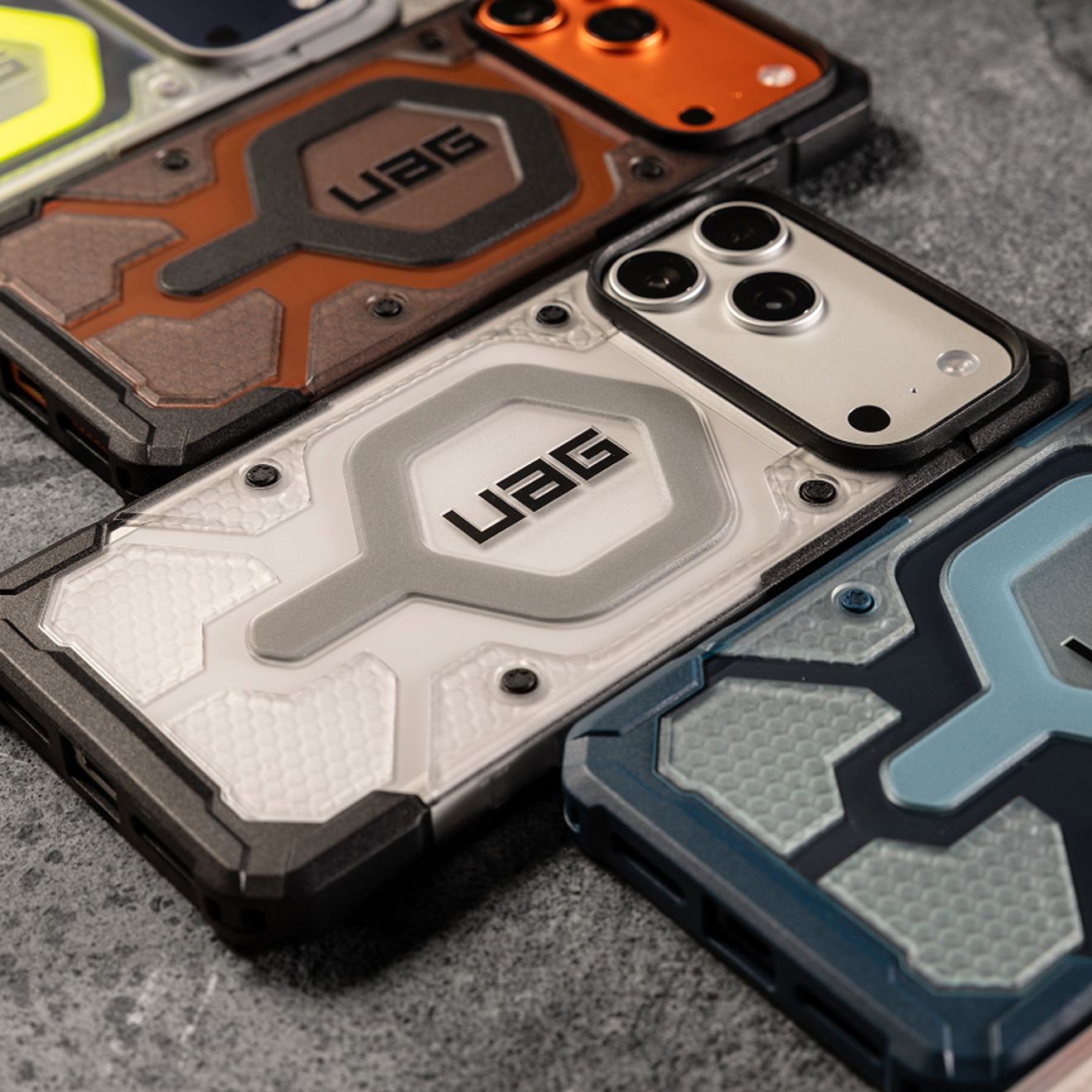 Cuatro iPhone Pro Max con fundas resistentes UAG en colores plata, naranja y azul están colocados uno al lado del otro, mostrando sus esquinas reforzadas, sus diseños con textura y la protección elevada para la cámara