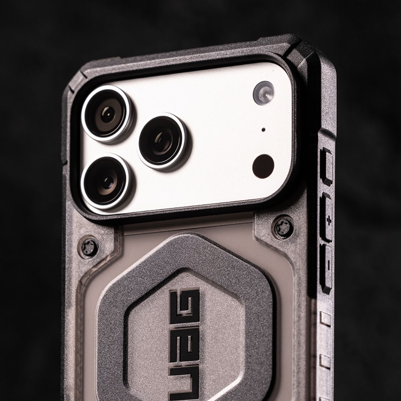 Primer plano del sistema de cámara de tres lentes de un iPhone Pro Max color plata, protegido por una resistente funda gris de UAG con bordes reforzados y botones laterales.