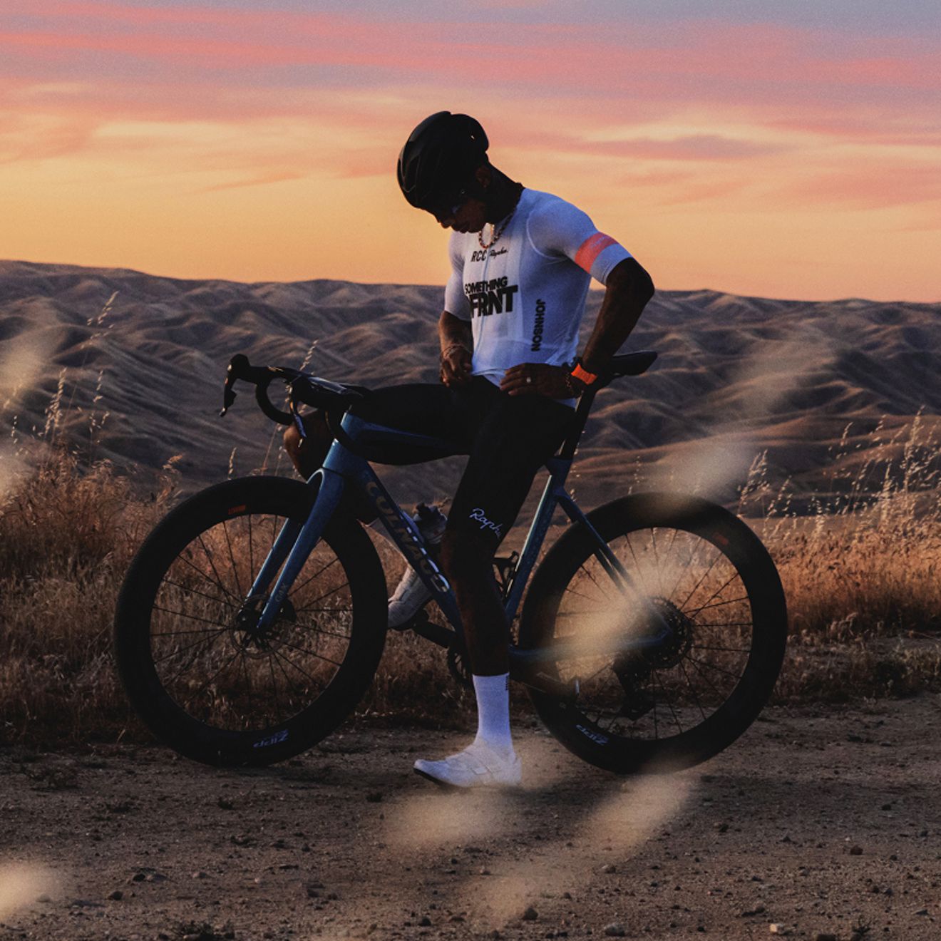 Un ciclista, con casco y ropa de ciclismo, está de pie junto a una bicicleta de carretera en un sendero de tierra, con colinas onduladas y una colorida puesta de sol al fondo.